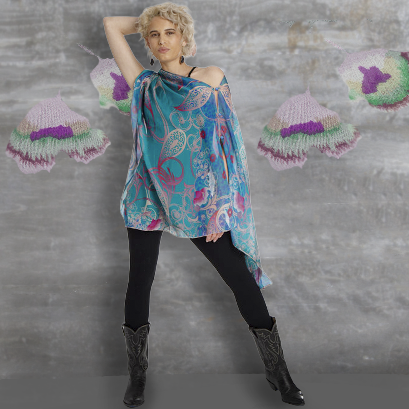 A woman models a draped, asymmetrical blue tunic top with a paisley and floral pattern in pink, purple, and white, styled with black leggings and black cowboy boots against a textured gray wall with hanging colorful knit fan decorations.
