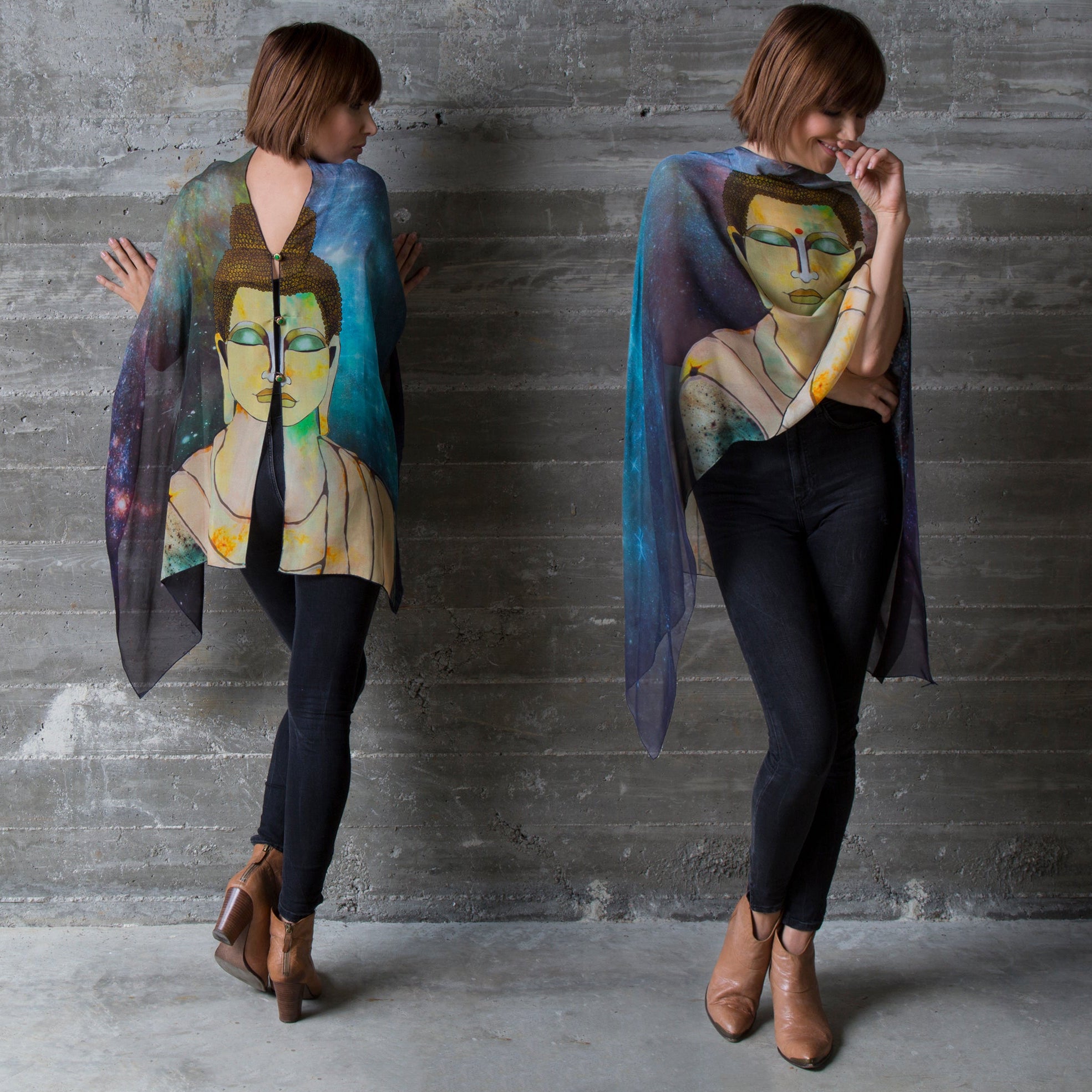 Two woman wearing a colorful LuLuWrap scarf with a painting of a Buddha face on it, standing against a gray concrete wall.